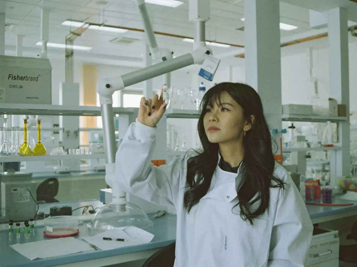 A woman in a lab coat and blue gloves carefully working with a pipette, holding two test tubes in her hands. She is in a laboratory setting, with a rack of test tubes visible in the foreground and a white wall in the background. The image is focused on her hands, emphasizing the scientific process.